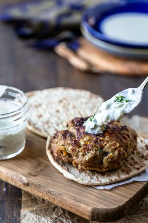 A spoon drizzling tzatziki on Greek chicken burger