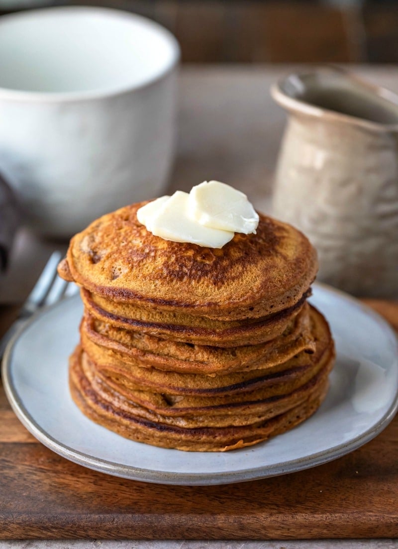 Stack of gingerbread pancakes on a blue plate next to a fork and a pitcher or syrup