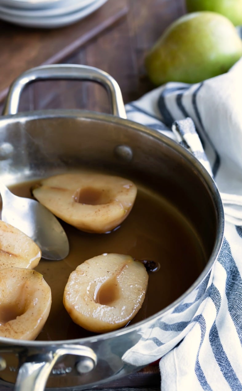 Cider poached pears in a silver skillet next to a striped dish towel