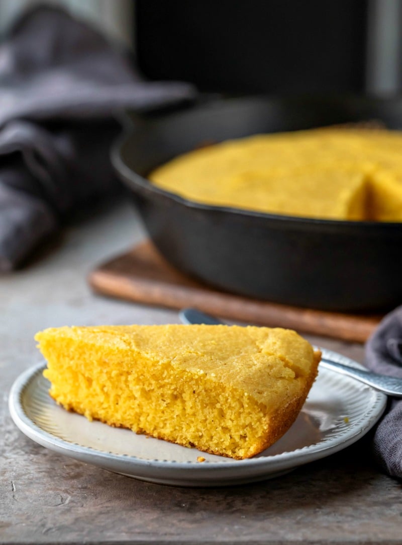 Slice of cornbread on a white plate next to a cast iron skillet