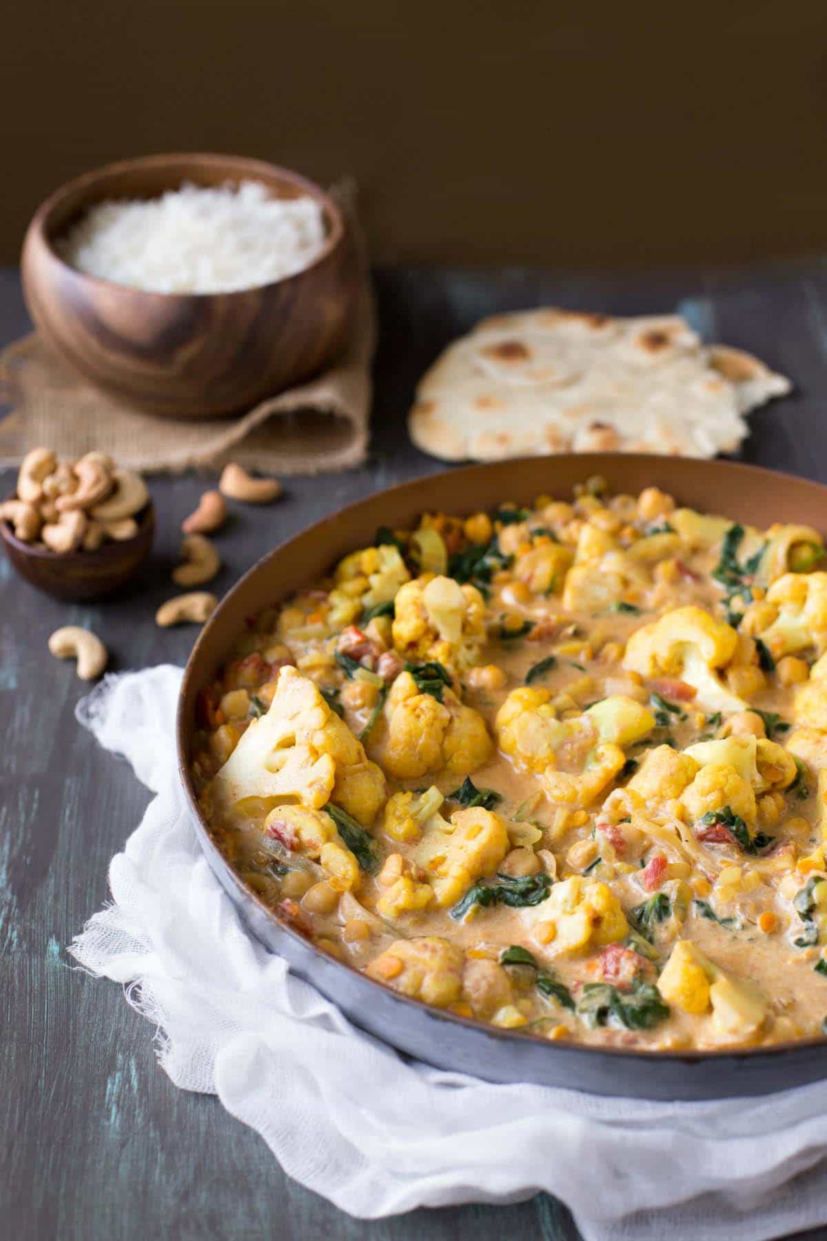 Vegetable Korma in a skillet next to a bowl of cashews and piece of naan