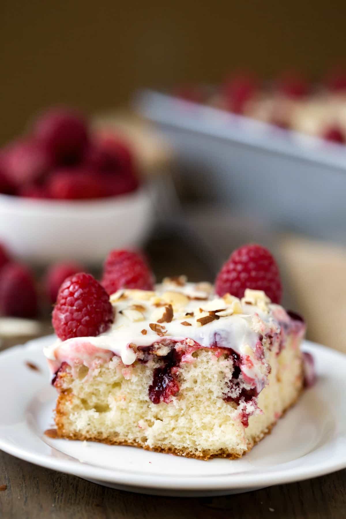 Slice of White Raspberry Poke Cake on a white plate next to a bowl of raspberries