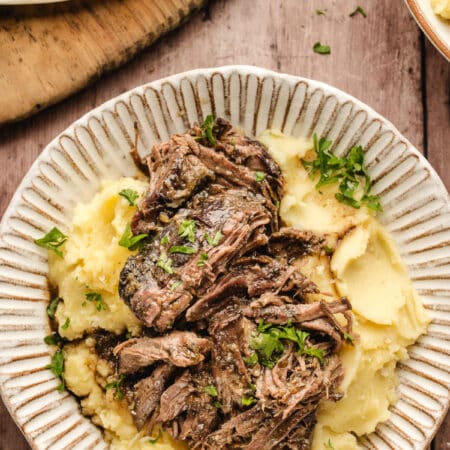 A dish of shredded Mississippi pot roast over mashed potatoes next to a dish of mashed potatoes.