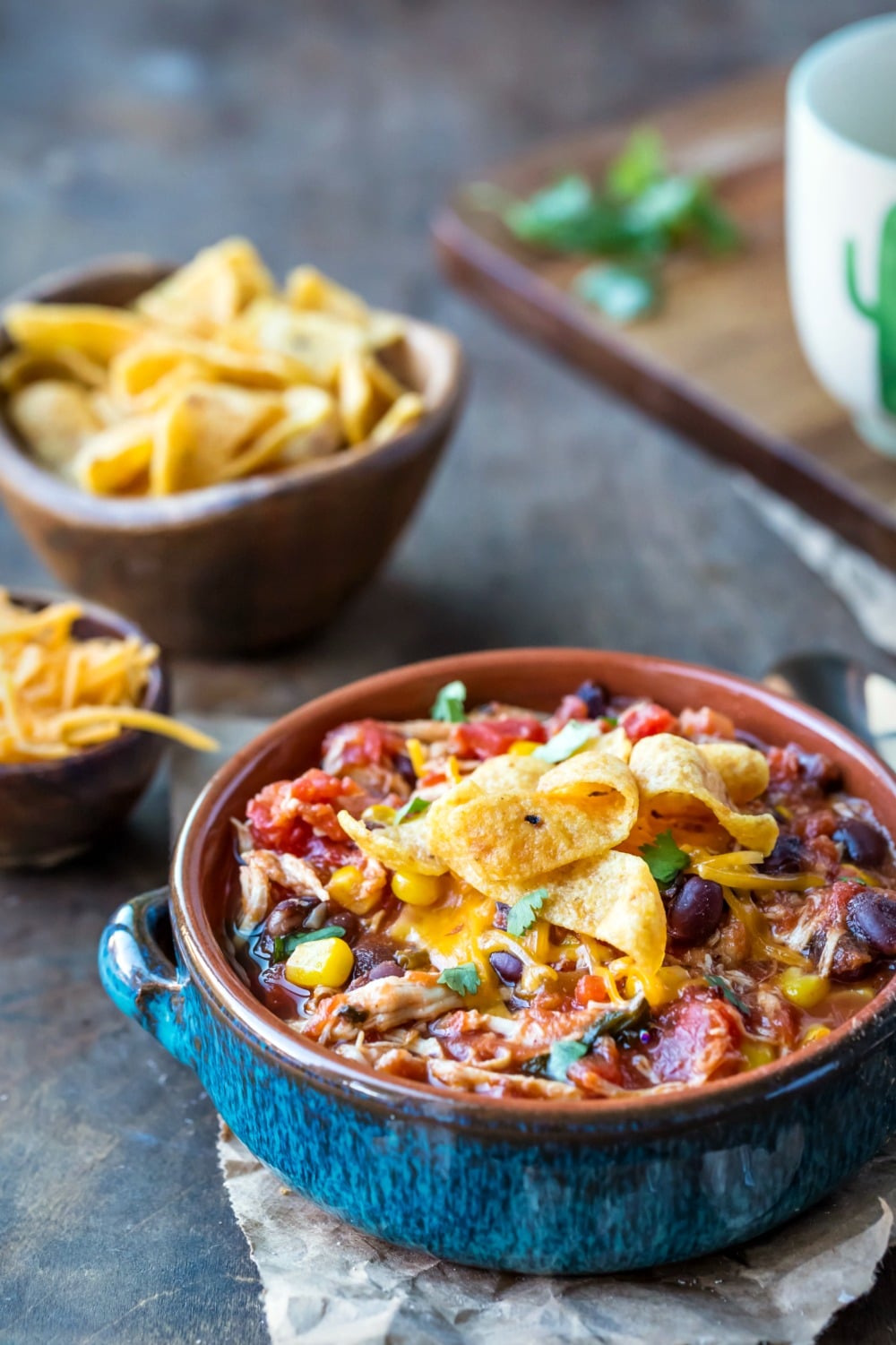 Slow Cooker Chicken Frito Chili in a blue dish next to shredded cheese and fritos