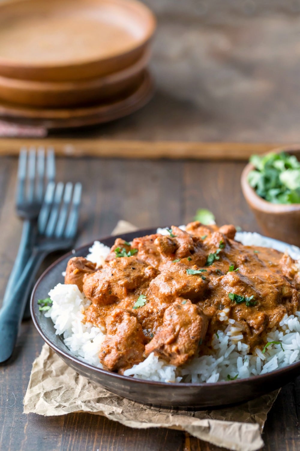 Indian butter chicken over rice in a dark dish next to a bowl of cilantro