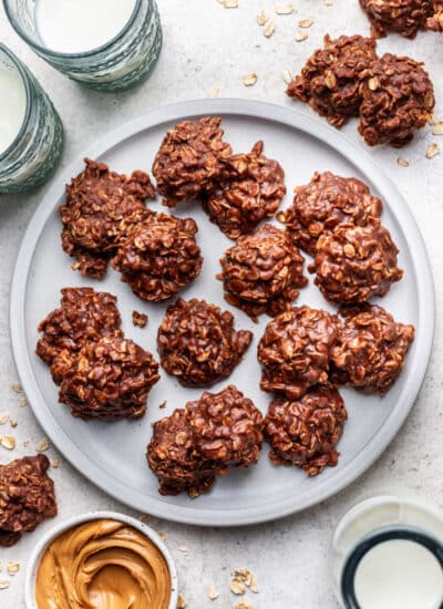 A plate of no bake cookies next to glasses of milk and a dish of peanut butter.