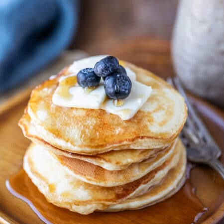 A stack of sour cream pancakes on a wooden plate topped with pats of butter and blueberries.