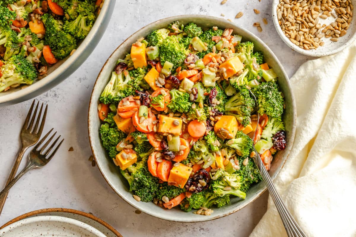 A dish of broccoli salad next to a stack of plates and forks. 