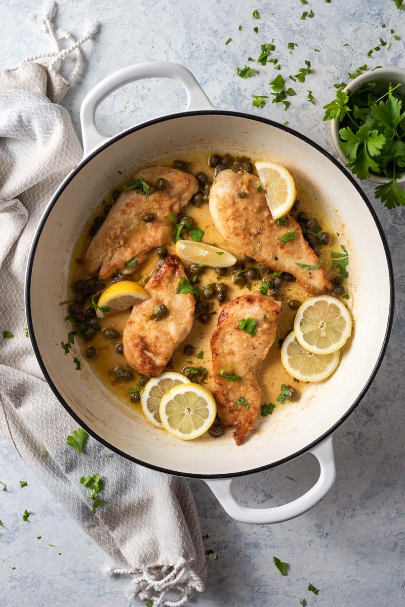 An overhead shot of chicken piccata in a white skillet with capers, lemon and parsley