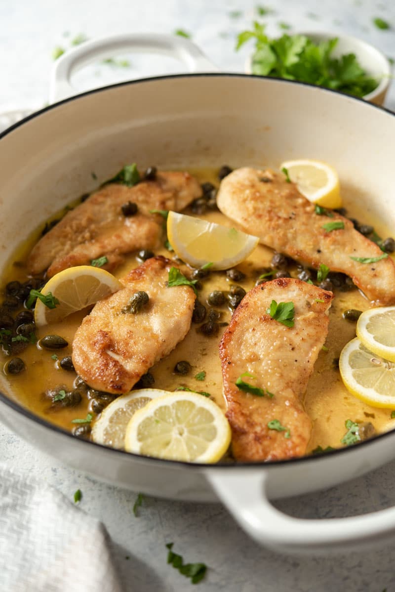 A side shot of chicken piccata in a white skillet with capers and slices of lemon