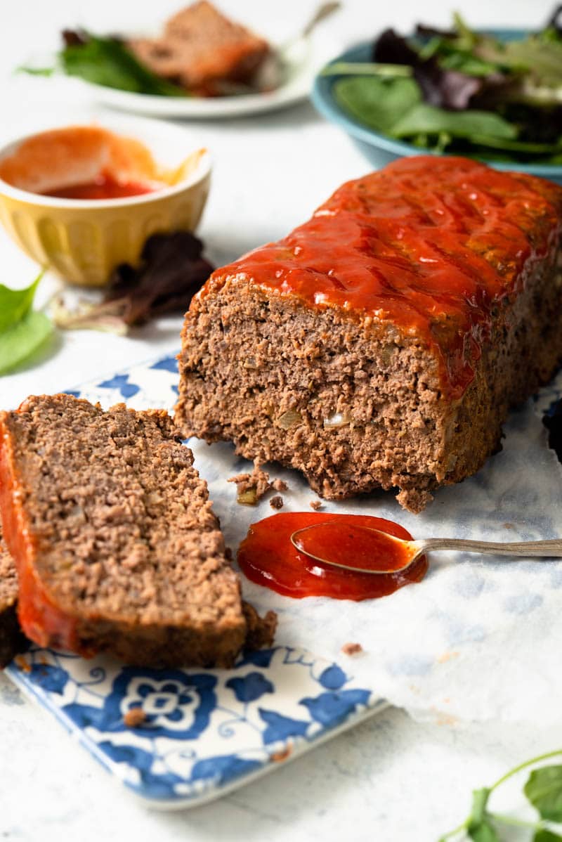 A side shot of meatloaf on a cutting board with some slices cut and chili sauce beside it