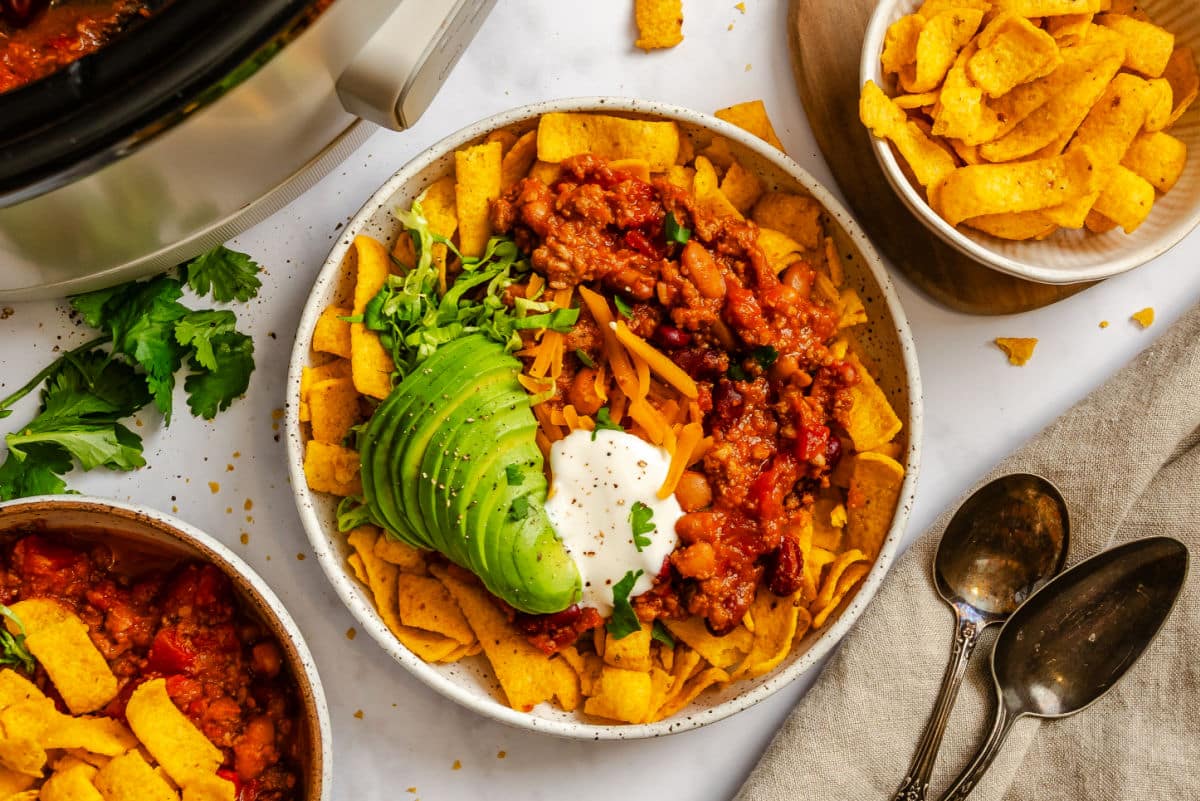 A dish of frito chili pie next to bowls of fritos and another bowl of chili pie.