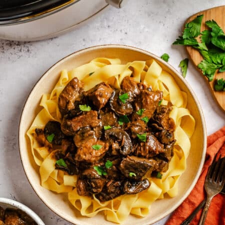 A plate of crock pot beef tips on egg noodles next to the crock pot.