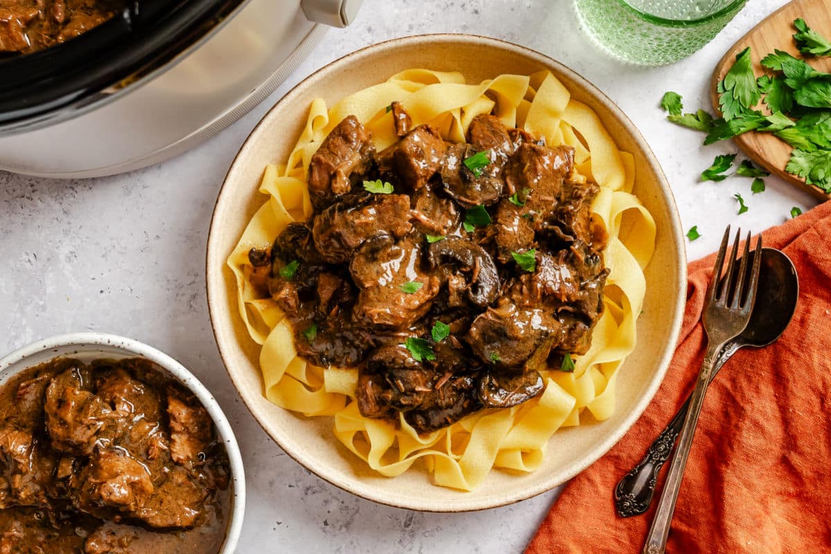 Crockpot Beef Tips A plate of crock pot beef tips on egg noodles next to chopped parsley and a glass of water.