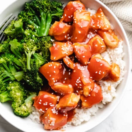 Orange chicken topped with sesame seeds in white dish next to steamed broccoli.