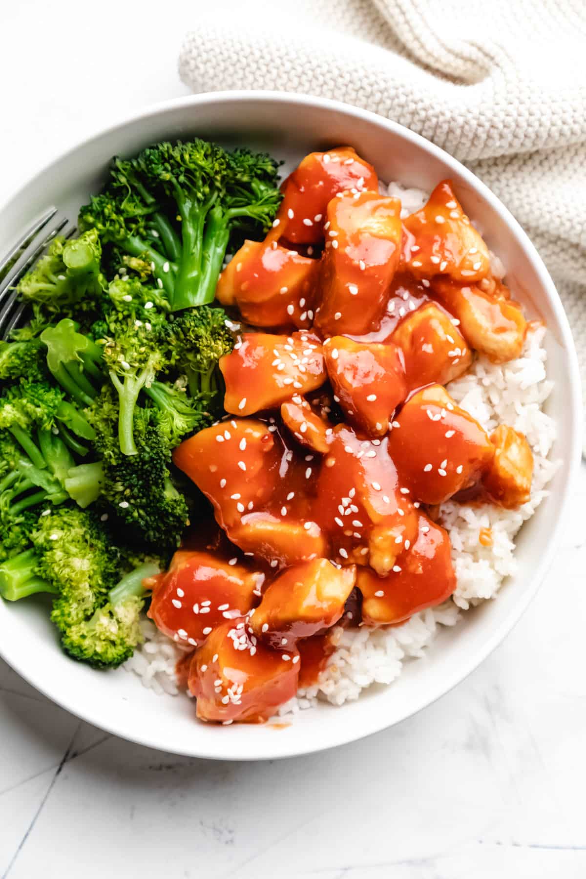 Orange chicken topped with sesame seeds in white dish next to steamed broccoli.