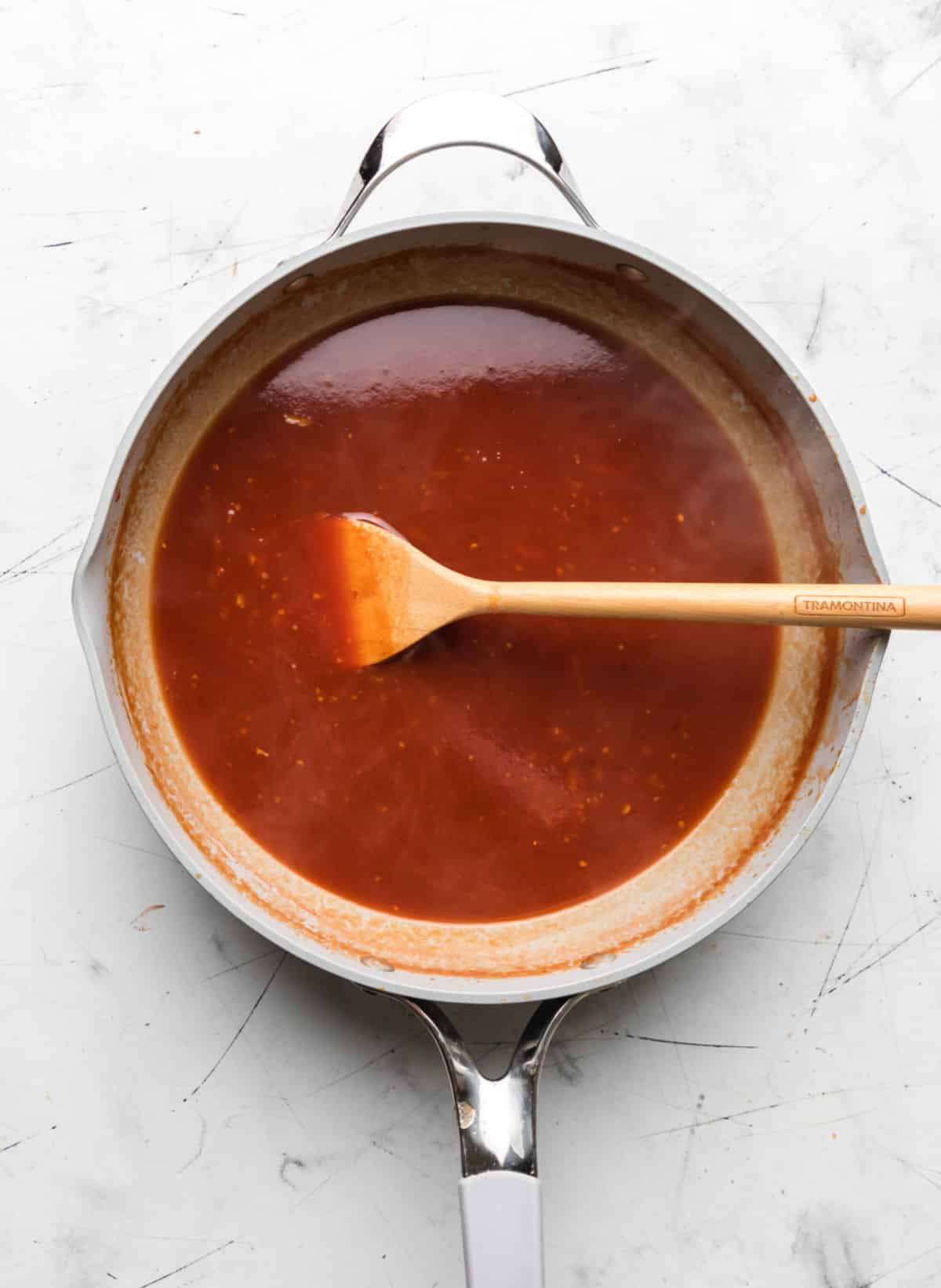 A wooden spoon in a skillet filled with orange chicken sauce. 