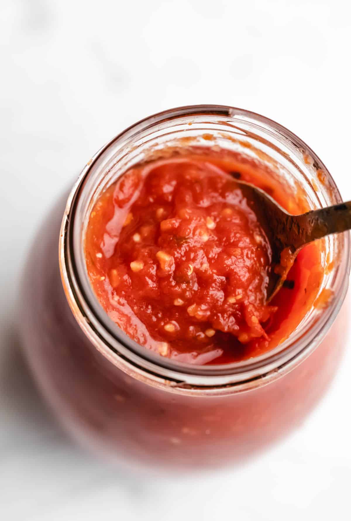A spoon scooping pizza sauce from a glass jar. 
