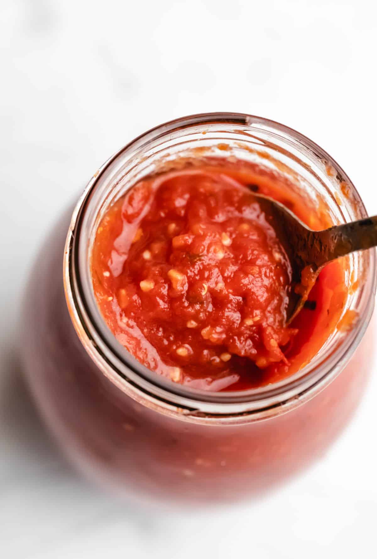 A spoon scooping pizza sauce from a glass jar.