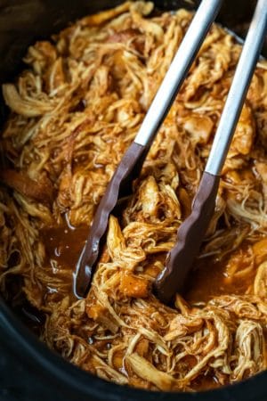 Tongs grabbing crock pot brown sugar bourbon bbq chicken in a crock pot