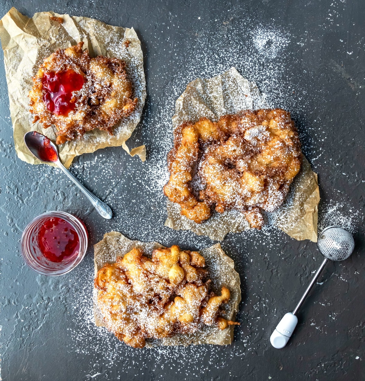 Three homemade funnel cakes next to a powdered sugar duster.