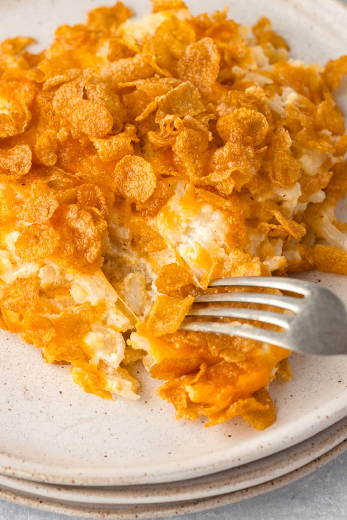 A fork taking a bite of cheesy funeral potatoes on a plate.