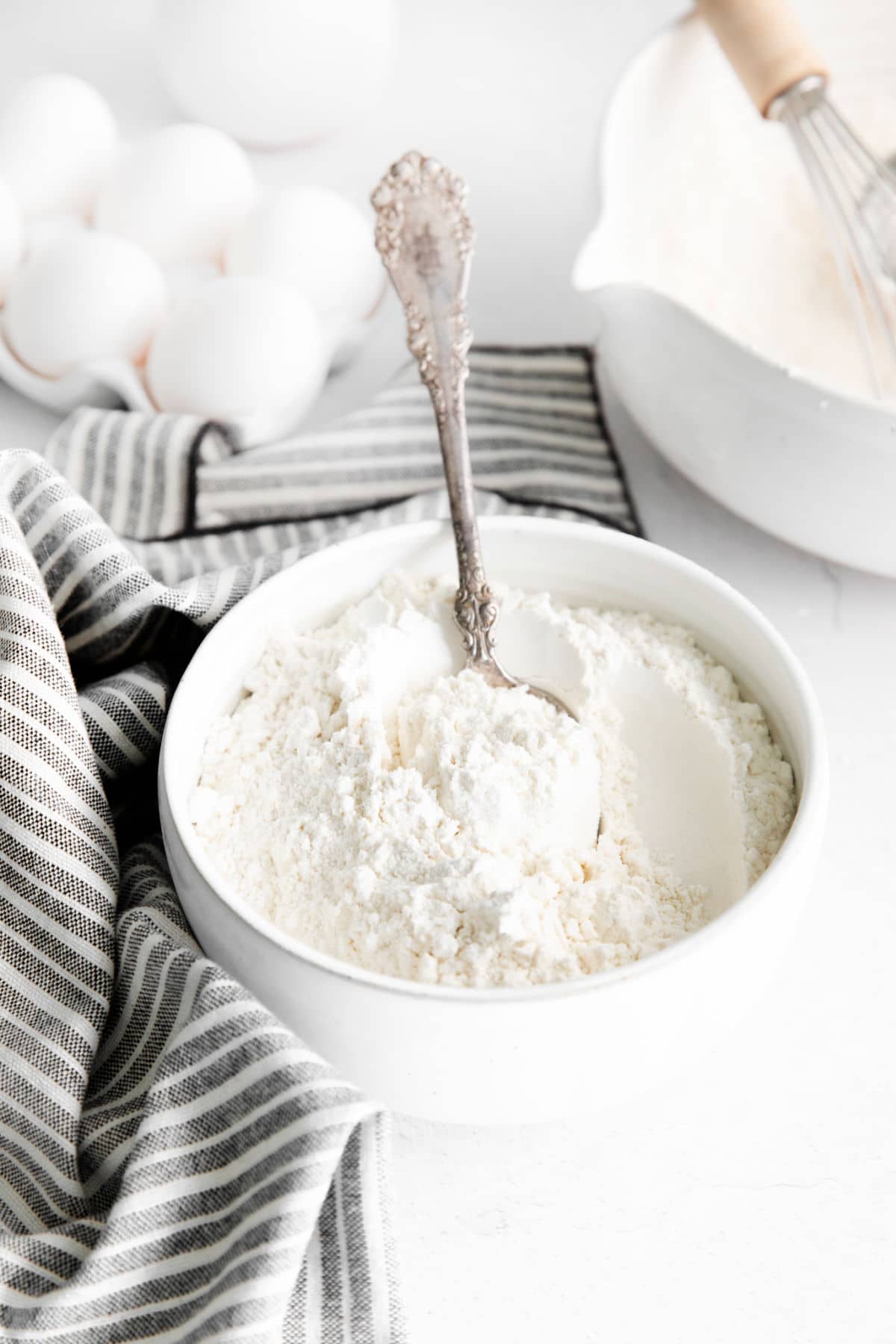 Bowl of cake flour next to eggs and a whisk.