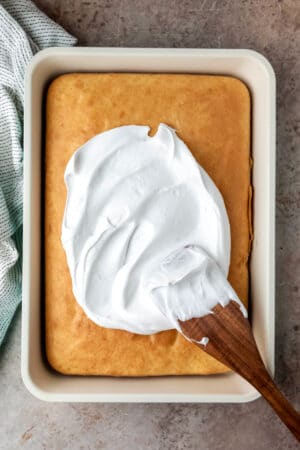 A wooden spatula spreading seven minute frosting onto a yellow cake.