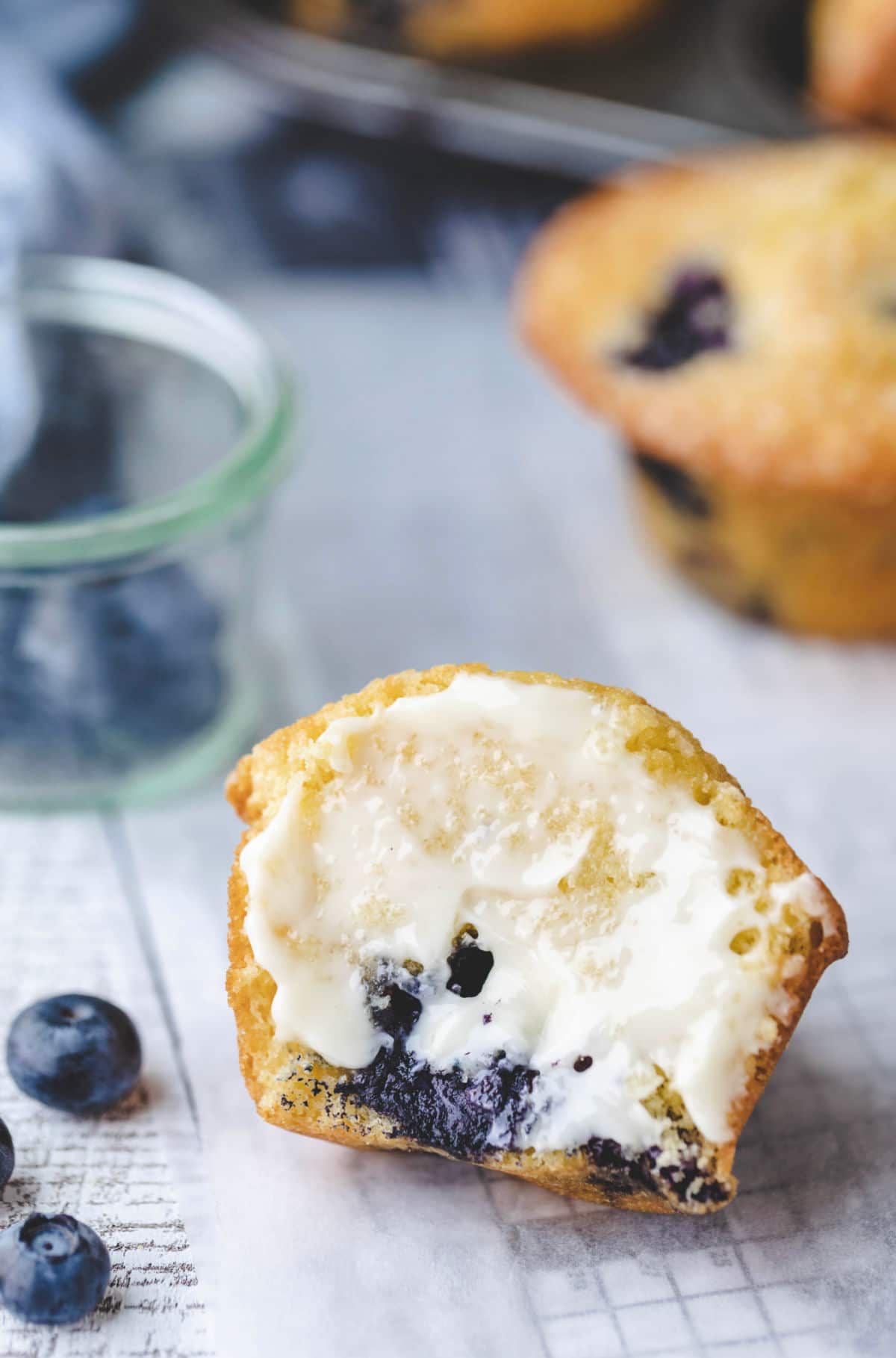 Half of a buttered blueberry muffin next to a glass jar with blueberries. 