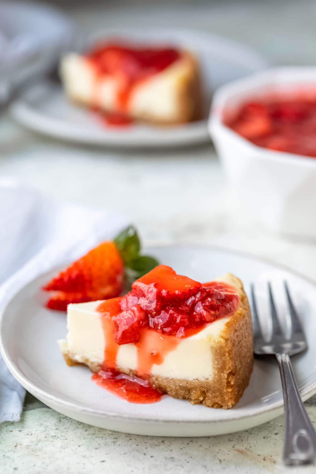 Slice of cheesecake covered with strawberry topping on a white plate. 