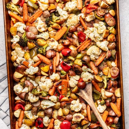 A wooden spoon resting in a sheet pan with roasted sausage (kielbasa) and veggies.