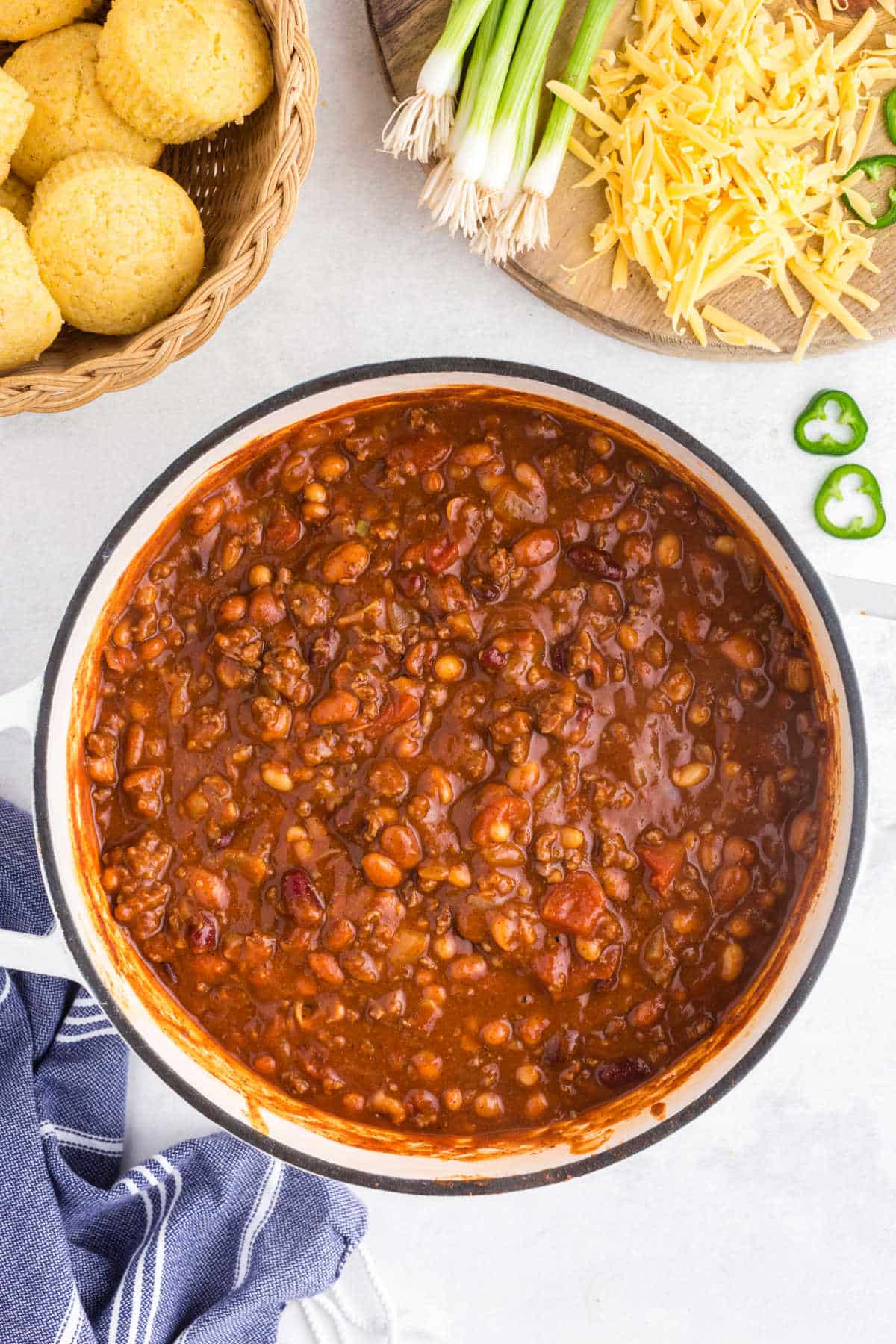 Pot of chili in a Dutch oven next to toppings and cornbread muffins.