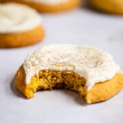 Frosted pumpkin cookie with a bite missing on a piece of white parchment paper.