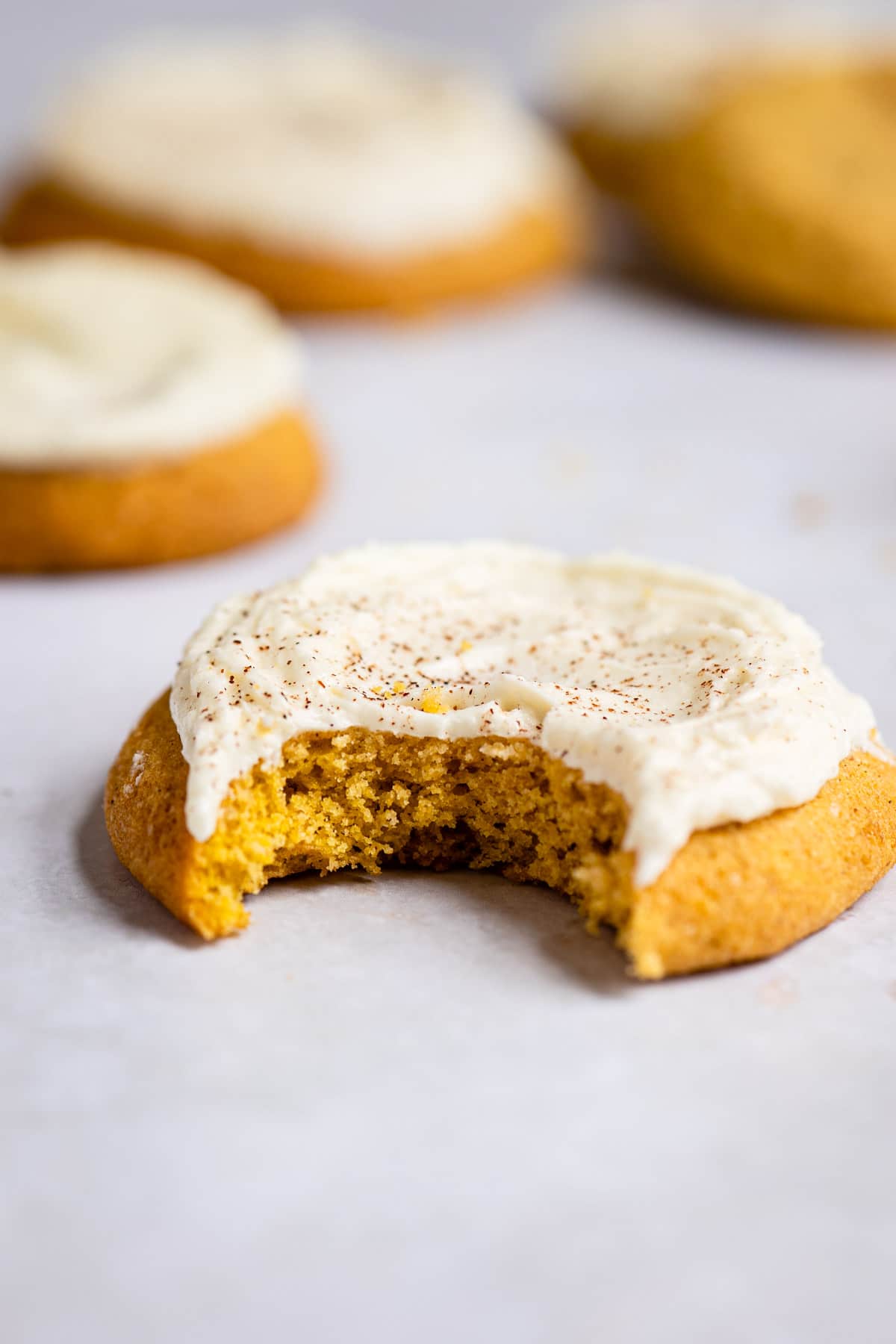 Frosted pumpkin cookie with a bite missing on a piece of white parchment paper. 