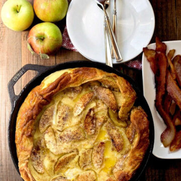 Caramelized apple German pancake next to apples and a plate of bacon.