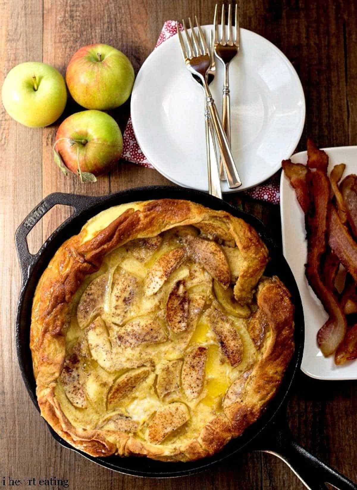 Caramelized apple German pancake next to apples and a plate of bacon.