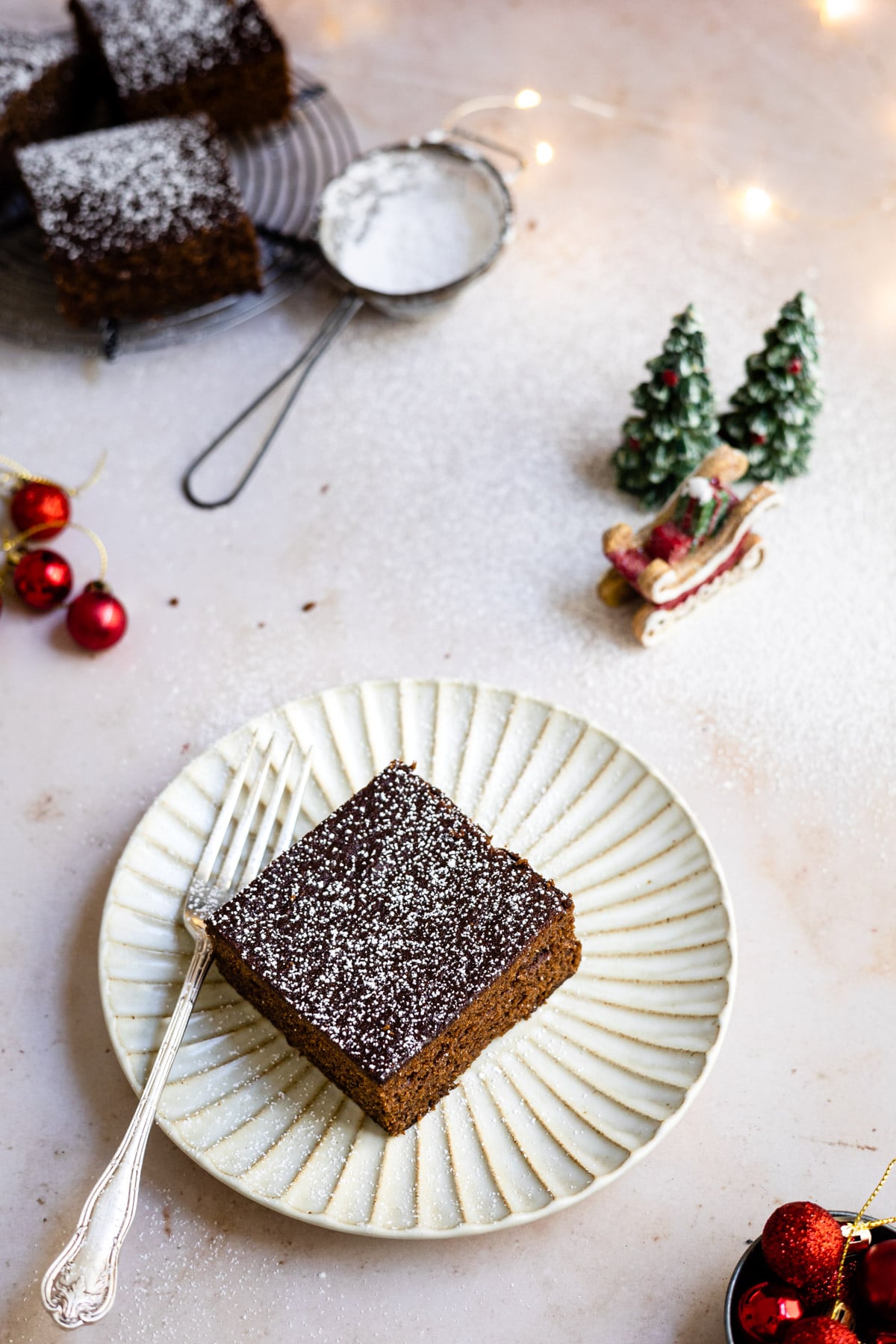 Piece of gingerbread cake on a white plate next to Christmas tree decorations.
