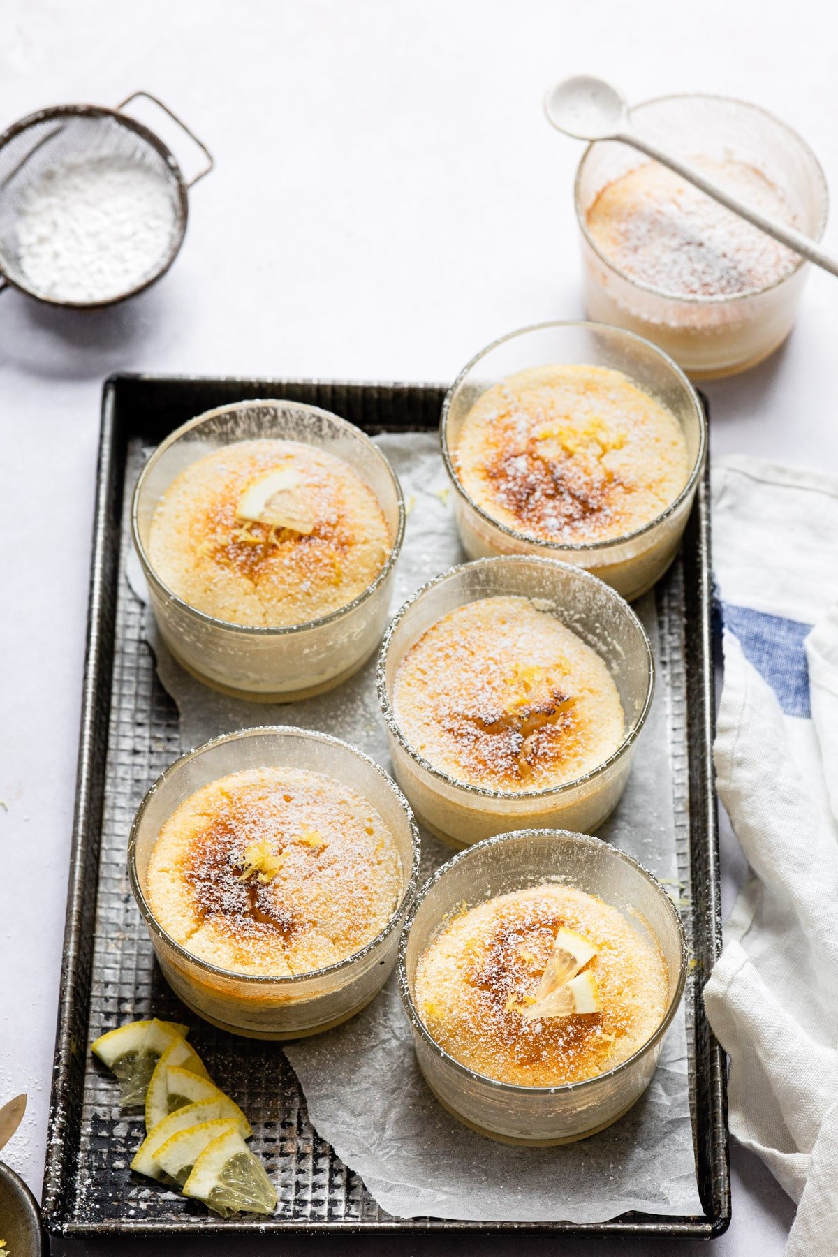 A tray with lemon pudding cakes on it next to a powdered sugar sifter. 