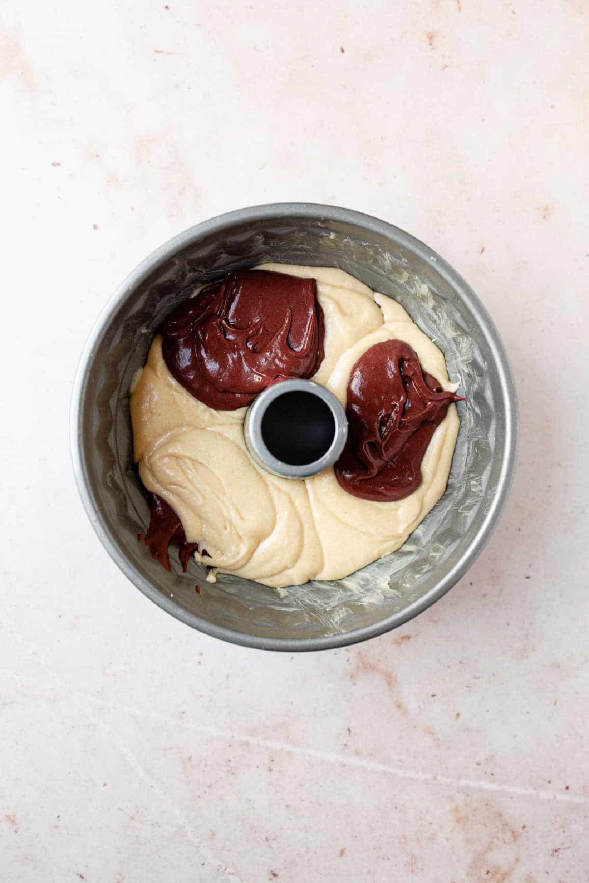 Two scoops of red velvet batter on plain cake batter in a bundt pan.