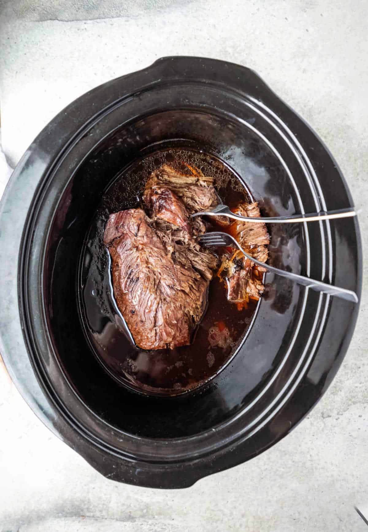 Two forks shredding a roast in a slow cooker. 