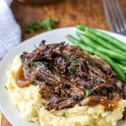 Crock pot balsamic beef on mashed potatoes next to green beans on a white plate.