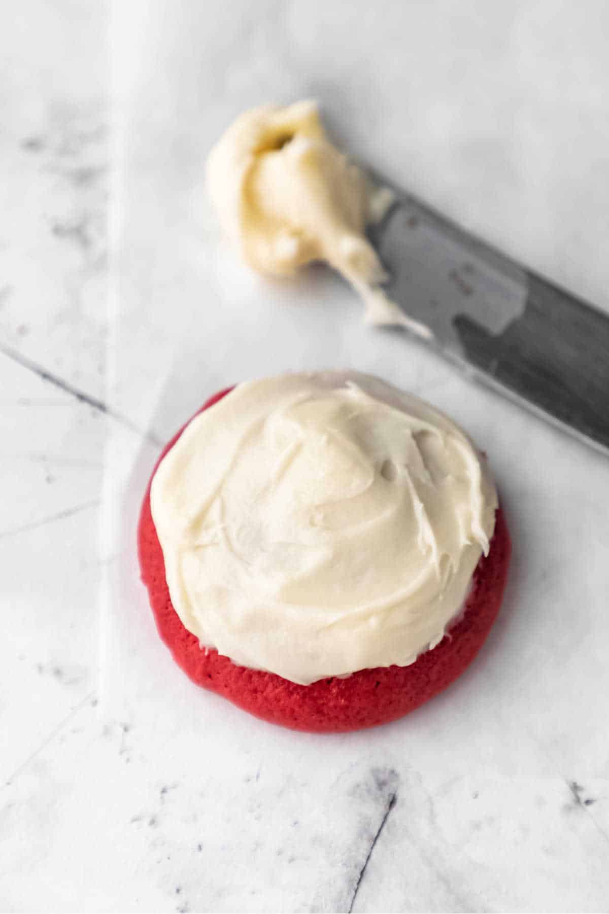A butter knife with cream cheese frosting on it next to a red velvet cookie.