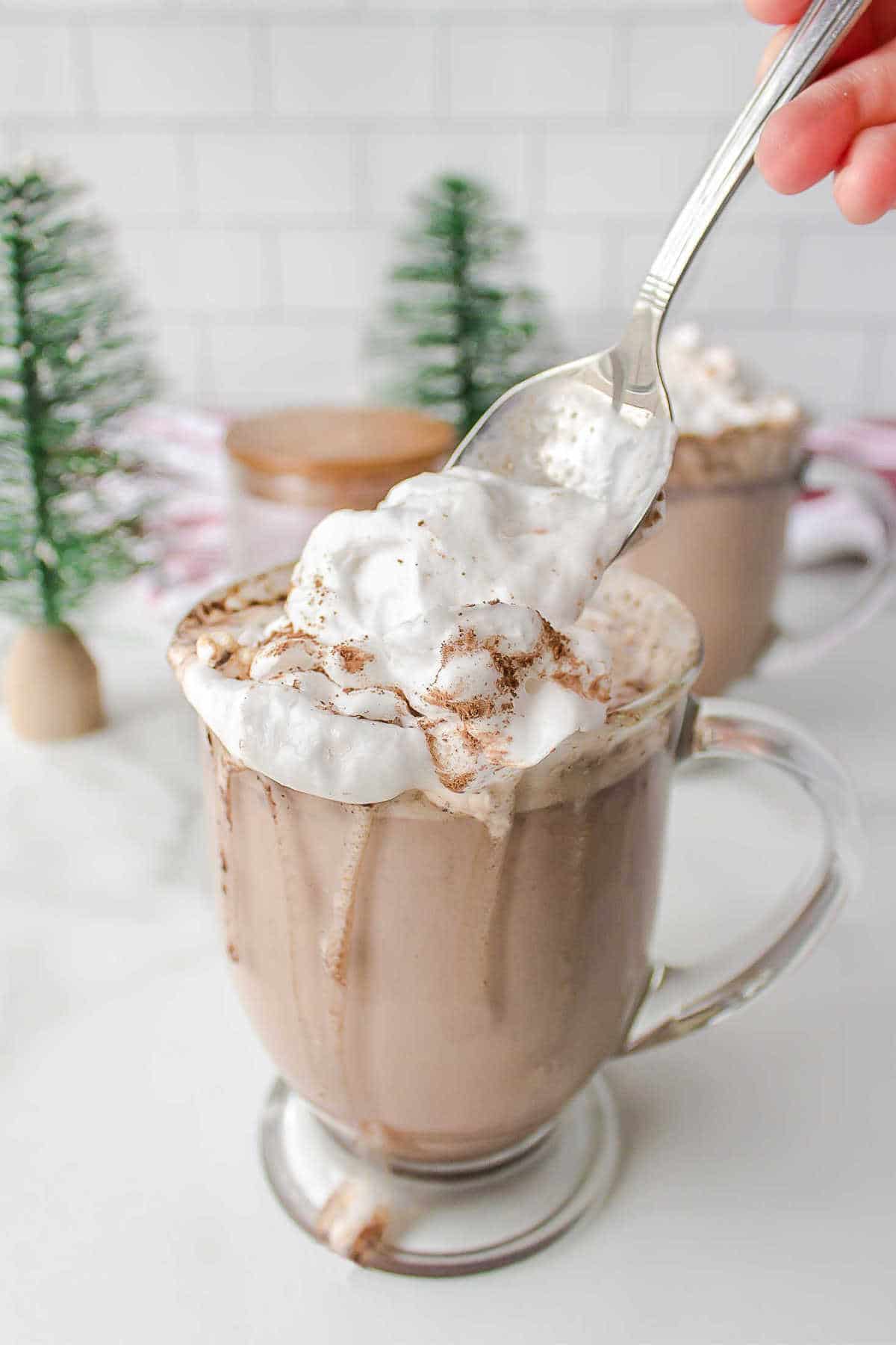 A spoon scooping whipped cream out of a glass mug of hot cocoa.