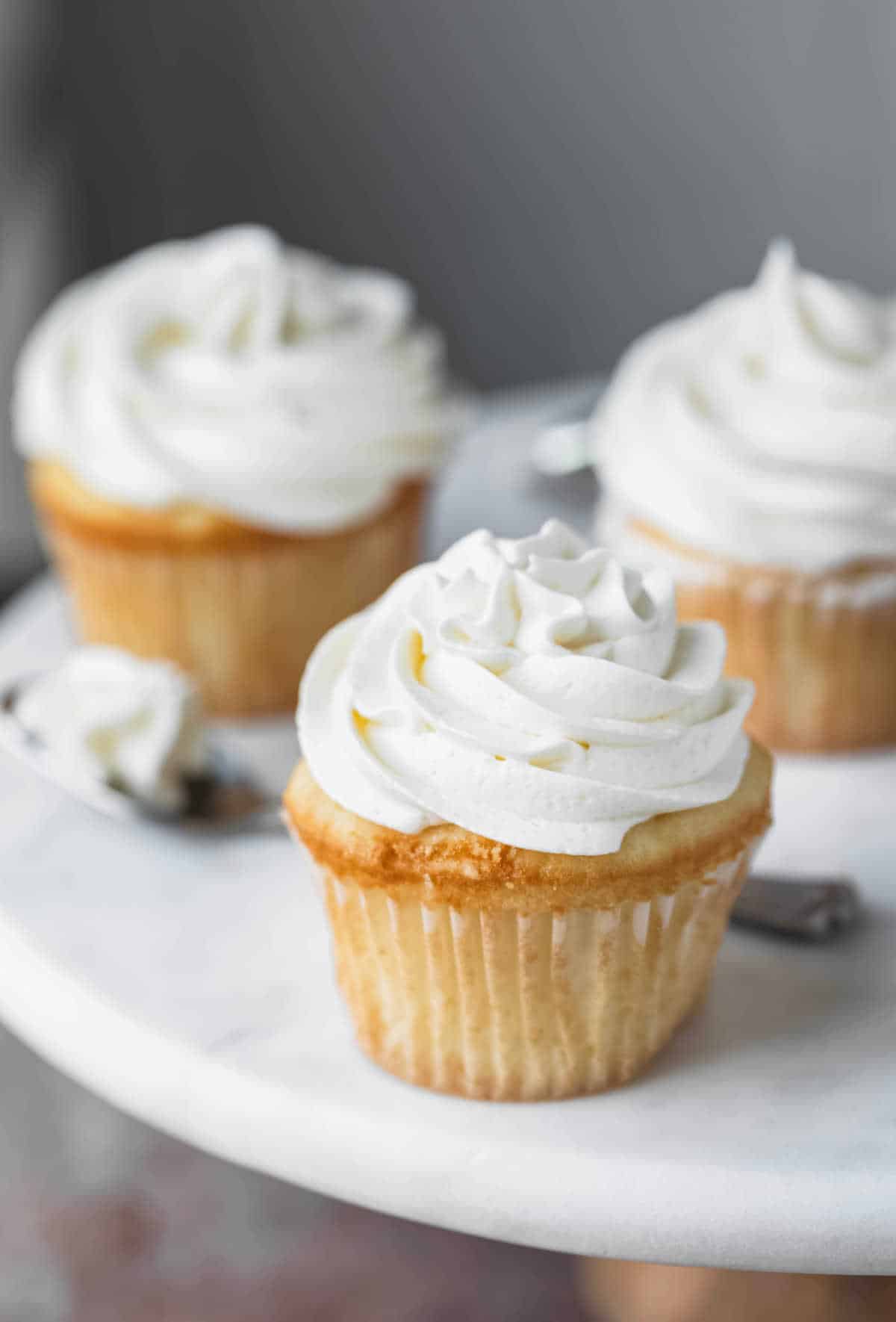 Vanilla Buttercream Frosting Three white cupcakes with vanilla buttercream frosting on a white marble cake stand.