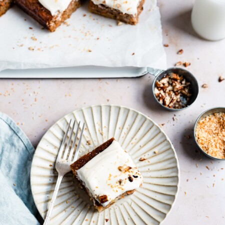 A piece of carrot cake on a white plate next to a tray of carrot cake slices.