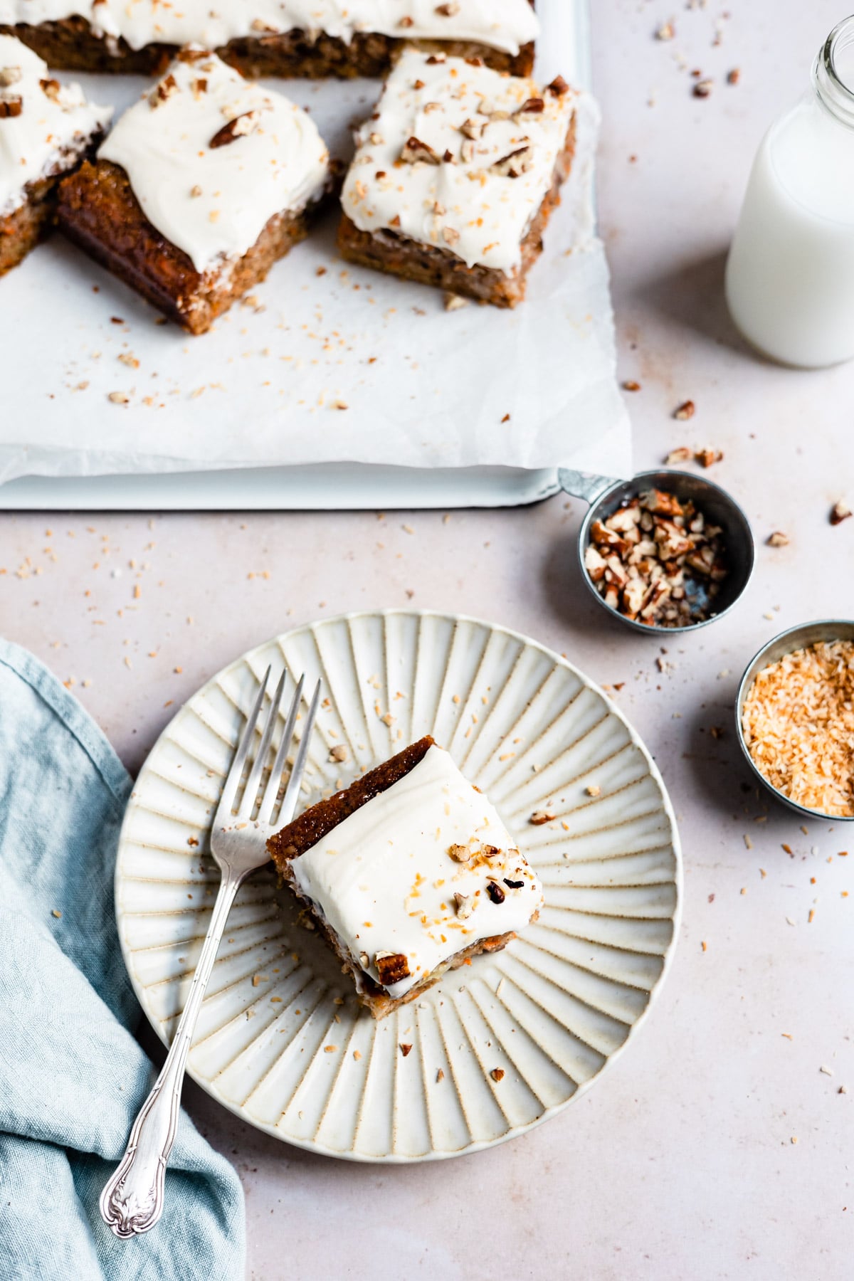 A piece of carrot cake on a white plate next to a tray of carrot cake slices. 