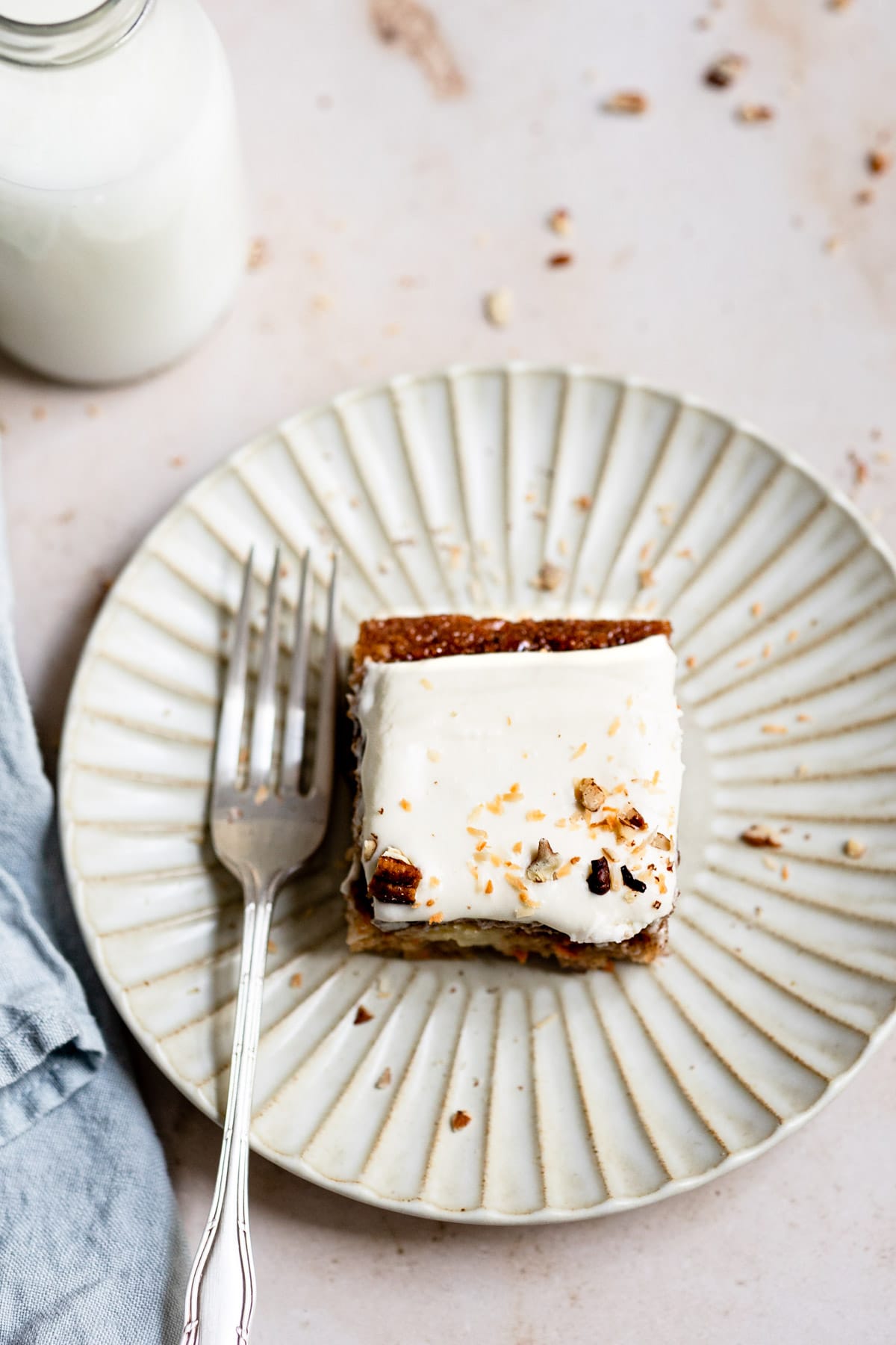 A slice of carrot cake on a white plate next to a glass of milk.