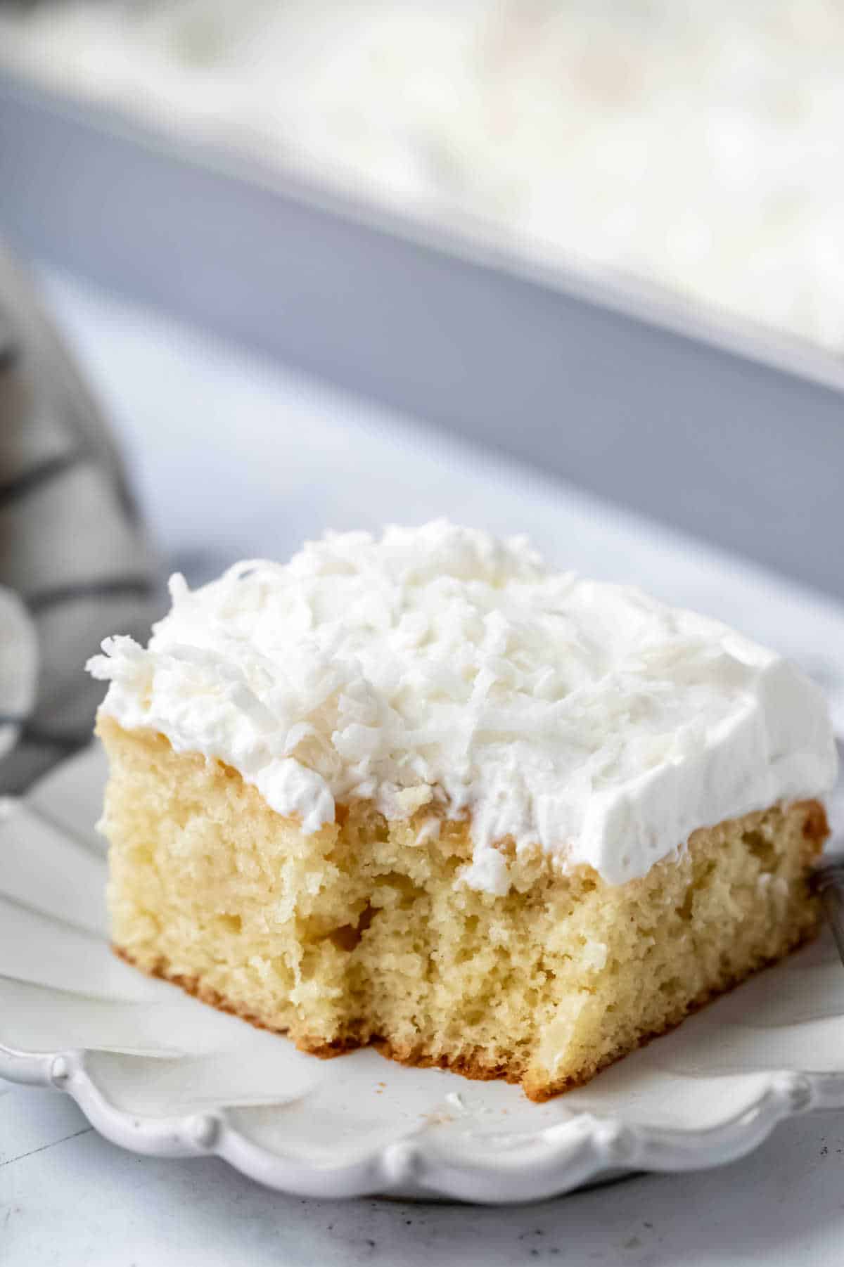 A slice of coconut poke cake on a plate with a bite missing. 