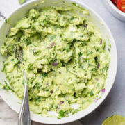 A dish of homemade guacamole next to cilantro tomatoes and lime.