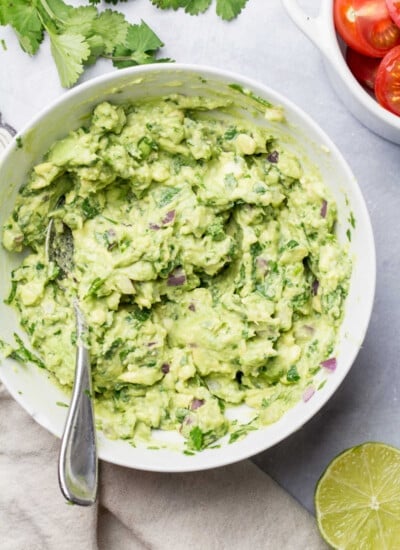 A dish of homemade guacamole next to cilantro tomatoes and lime.